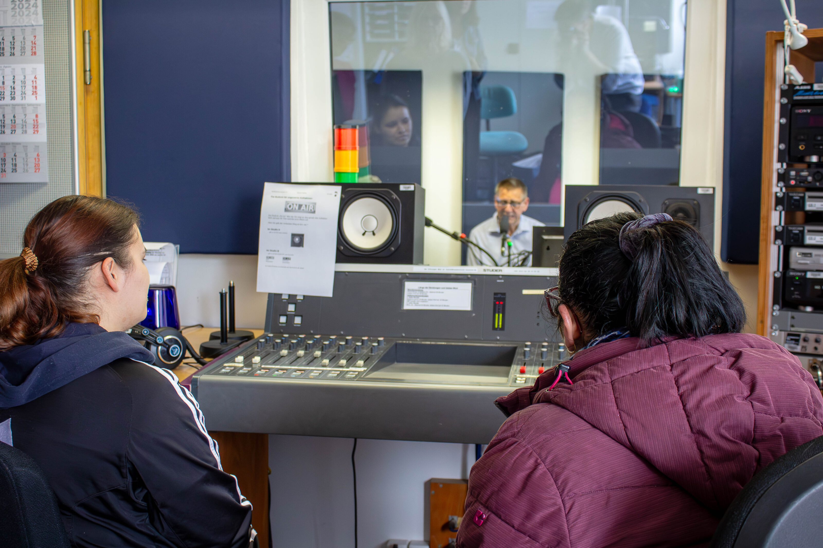 Zwei Frauen sitzen vor einem Mischpult und gucken in die Radiokabine, in der ein Mann ins Mikrophon spricht