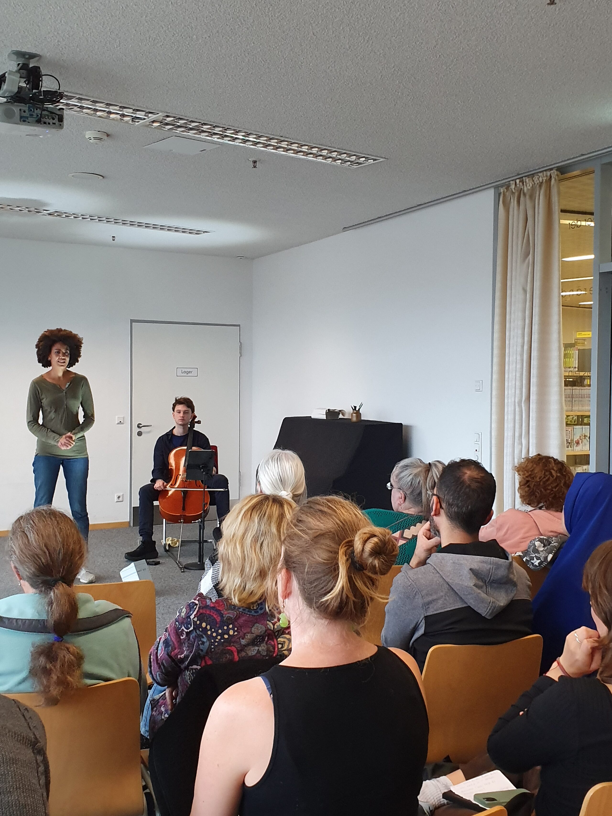 Publikum blickt auf eine Schauspielerin und Cello-Spieler neben Klavier in einer Bibliothek